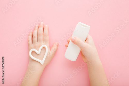 White heart shape on baby girl hand. Light pink table background. Pastel color. Hand holding little tube of moisturizing cream. Closeup. Point of view shot. Care about child hands skin. Top down view.