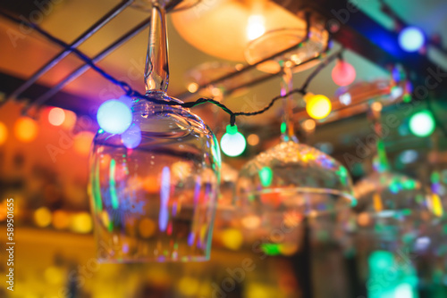 Bar interior details, glasses for wine and different alcohol cocktail drinks hang over bar counter with alcohol bottles and bartender in the background, nightlife club with backlit illumination