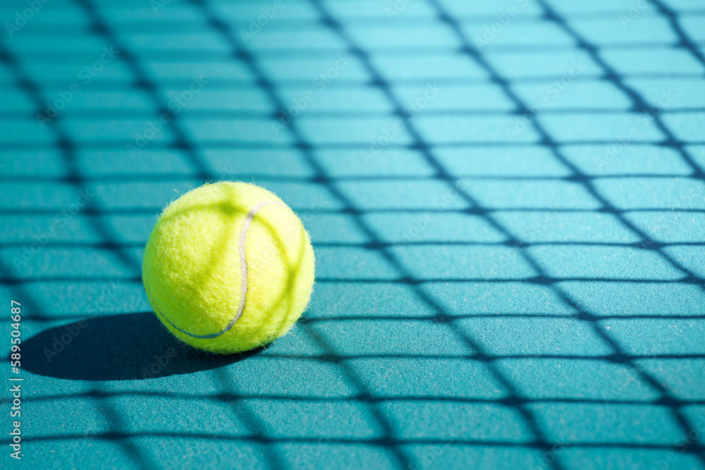 close-up of a tennis ball lying on a hard blue court in the shadow of a tennis net