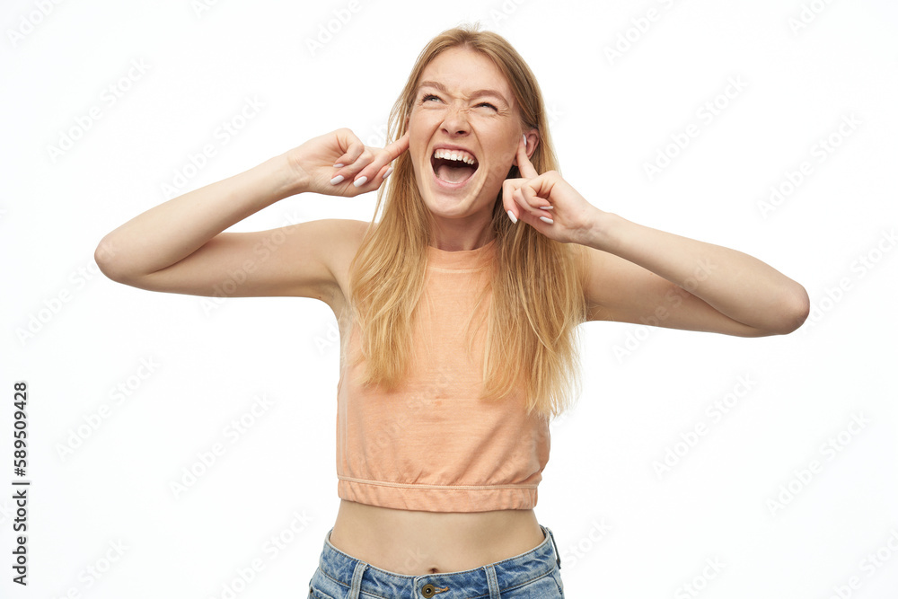 Indoor studio portrait of young ginger female with freckles closed her ears and screaming. isolated over background Indoor studio portrait of young ginger female with freckles closed her ears and screaming. isolated over background