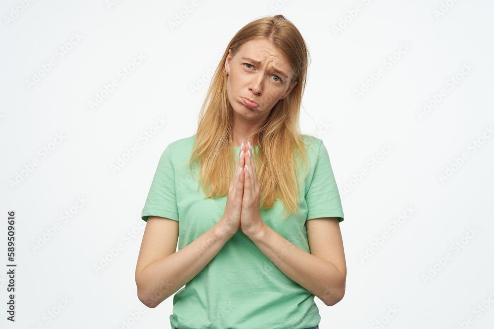 Indoor studio portrait of young ginger female with freckles press palms together, curving her lip and praying isolated over background Indoor studio portrait of young ginger female with freckles press palms together, curving her lip and praying isolated over background