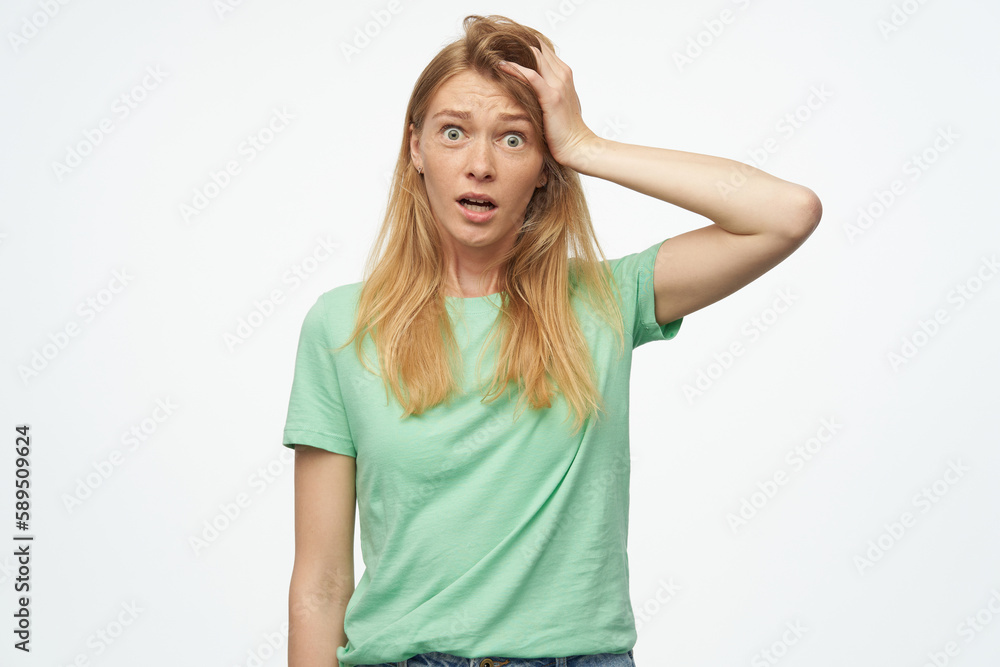 Indoor studio portrait of young ginger female with freckles feels confused, touching her had isolated over background Indoor studio portrait of young ginger female with freckles feels confused, touching her had isolated over background
