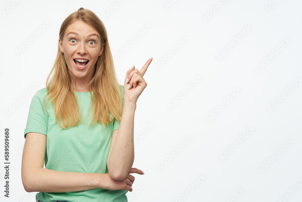 Indoor studio portrait of young ginger female with freckles point with a finger aside at copy space isolated over background