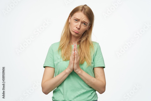 Indoor studio portrait of young ginger female with freckles press palms together, curving her lip and praying isolated over background