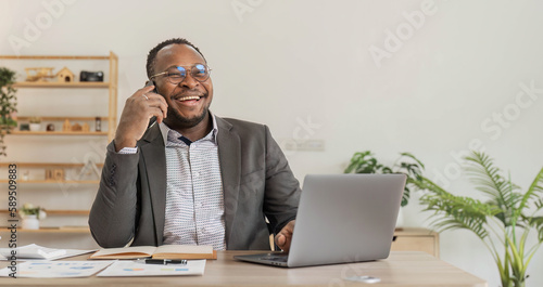 Portrait of handsome African black young business man working on laptop and talking phone at office desk.