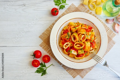 Italian Traditional Dish"Calamarata con calamari",calamarata pasta with squid,cherry tomatoes,olive oil,white wine,garlics,parsley,salt and peppers on plate with white wood background.Top view