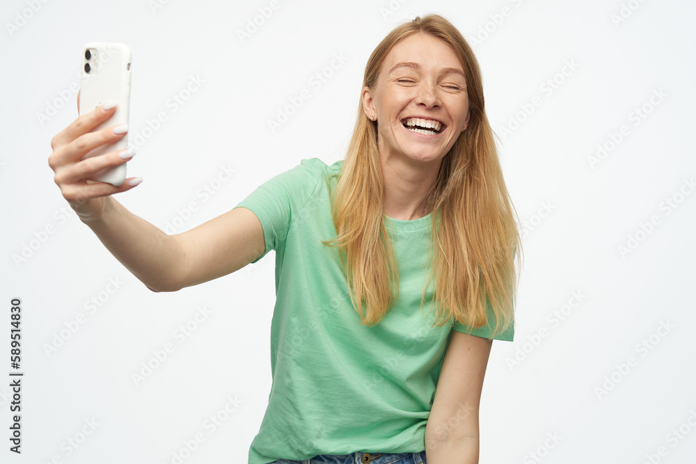 Indoor studio portrait of young ginger female with freckles making selfie and smiles broadly isolated over background Indoor studio portrait of young ginger female with freckles making selfie and smiles broadly isolated over background