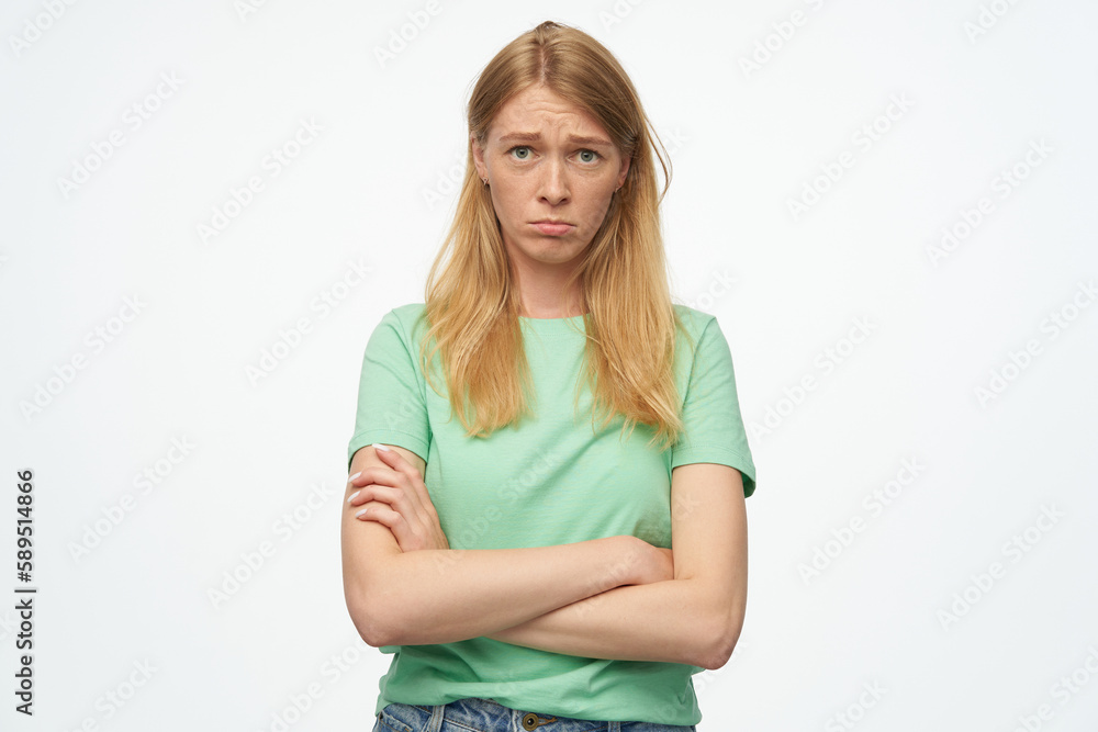 Indoor studio portrait of young ginger female with freckles looking into camera, keeps hands folded with sad facial expression, isolated over background Indoor studio portrait of young ginger female with freckles looking into camera, keeps hands folded with sad facial expression, isolated over background