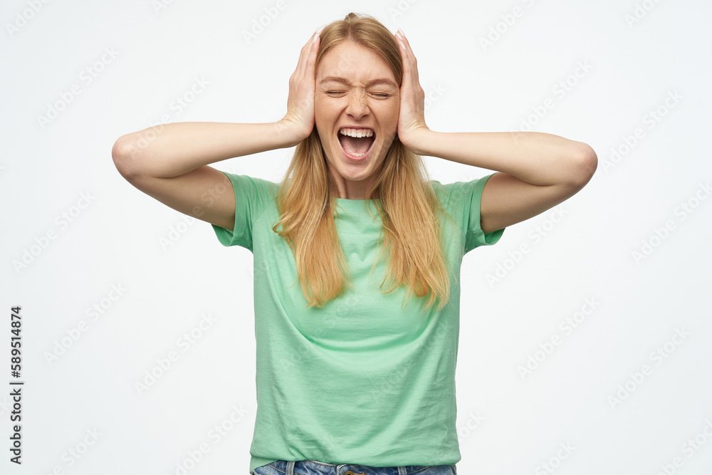 Indoor studio portrait of young ginger female with freckles closed her ears with palms and screaming, isolated over background Indoor studio portrait of young ginger female with freckles closed her ears with palms and screaming, isolated over background