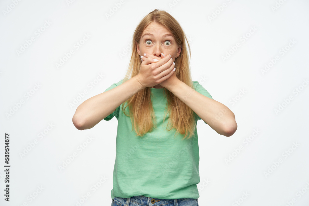Indoor studio portrait of young ginger female with freckles closed her mouth with palms, isolated over background Indoor studio portrait of young ginger female with freckles closed her mouth with palms, isolated over background