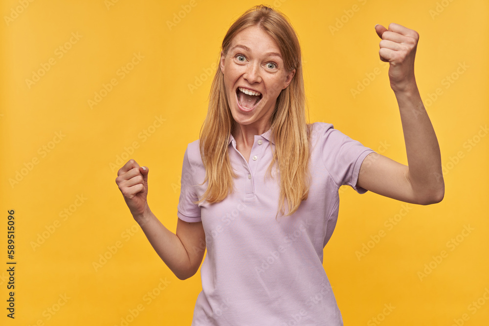 Indoor studio portrait of young ginger female with freckles celebrating victory, isolated over yellow orange background Indoor studio portrait of young ginger female with freckles celebrating victory, isolated over yellow orange background