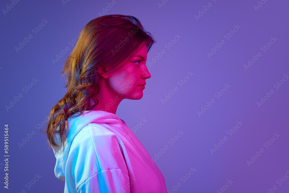 Side view portrait of young pretty girl looking straight with serious facial expression against gradient blue purple studio background in neon light. Concept of emotions, youth, lifestyle Side view portrait of young pretty girl looking straight with serious facial expression against gradient blue purple studio background in neon light. Concept of emotions, youth, lifestyle