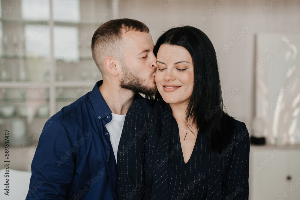 Young adult caucasian man home kissing wife, happy anniversary. Beardy eyes closed American guy embracing wife at hotel room on honeymoon. Successful businesswoman enjoys holiday with husband.