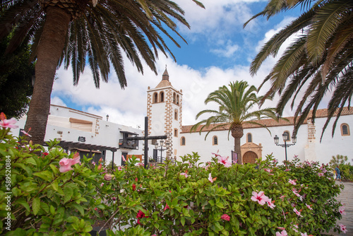 Vista panorámica de la iglesia Santa María de Betancuria en Fuerteventura, Islas Canarias. Impresionante arquitectura blanca rodeada de mucha vegetación, palmeras y flores rosas en un día nublado.