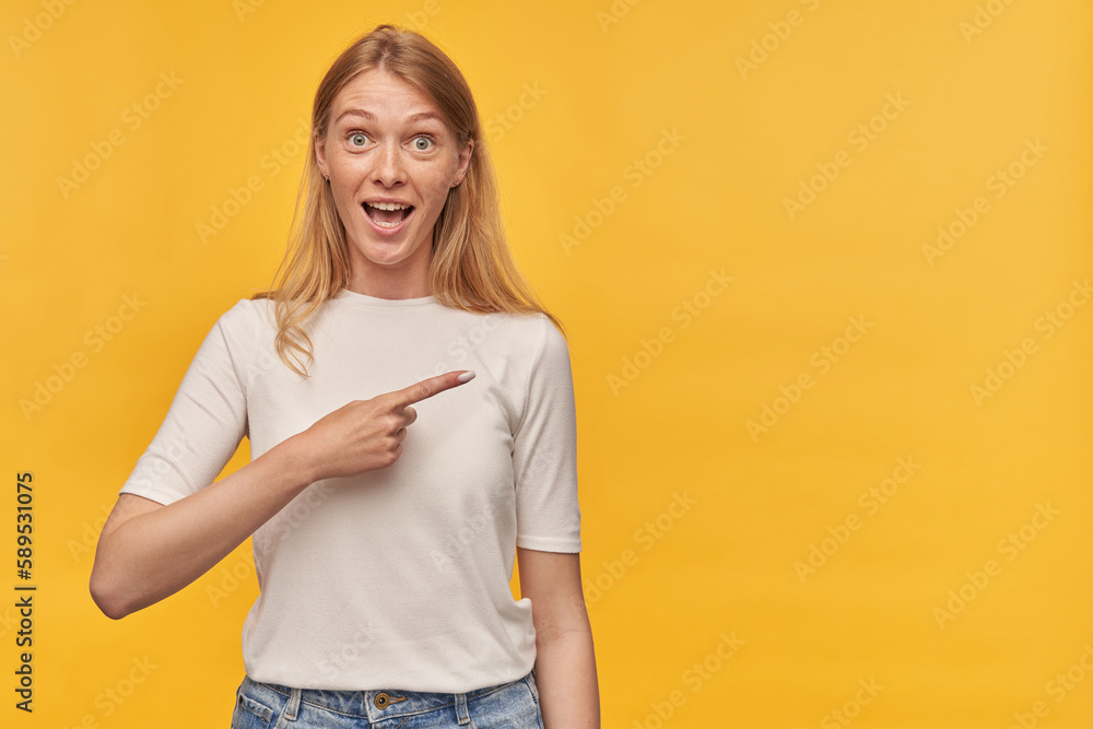 Indoor studio portrait of young ginger female with freckles points with a finger aside, isolated over yellow orange background Indoor studio portrait of young ginger female with freckles points with a finger aside, isolated over yellow orange background