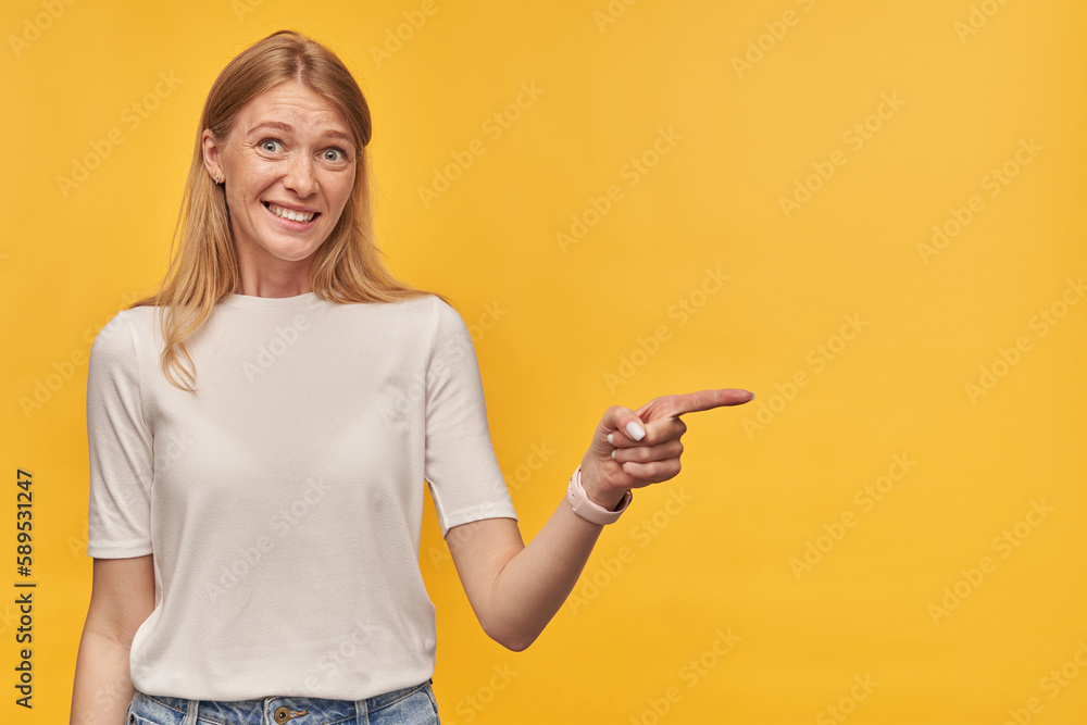 Fototapeta premium Indoor studio portrait of young ginger female with freckles points with a finger aside, isolated over yellow orange background
