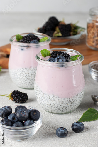 Chia pudding with chocolate muesli, fresh blueberry, blackberry and yogurt in two glass jars on light gray table. Healthy breakfast concept