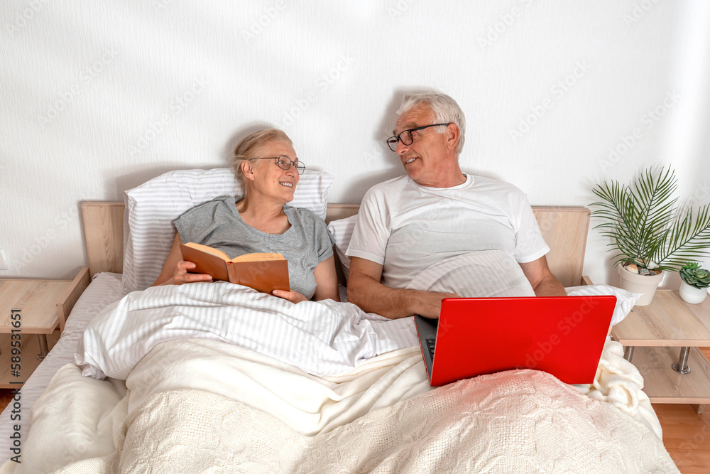 Senior couple spending leisure time in bed. A woman reads a book, a man works with a laptop while lying in the bedroom