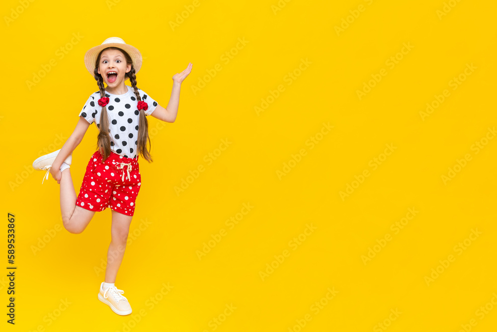 A beautiful girl in shorts points to your advertisement. The child points to your ad. A little lady in a hat and shorts on a yellow isolated background. Copy space. Banner A beautiful girl in shorts points to your advertisement. The child points to your ad. A little lady in a hat and shorts on a yellow isolated background. Copy space. Banner