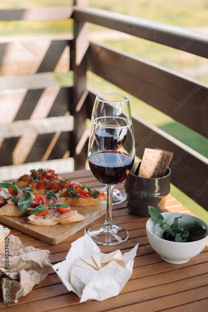 Glasses of red wine, bruschetta with tomato and basil, cheese, olives. Table setting for relaxing at summer terrace on sunny day.