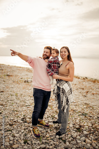 Young happy family spending their vacation on Italian Garda lake coast, bearded daddy pointing with finger, smiling mom, their little toddler girl in red checkered dress  © Natalia