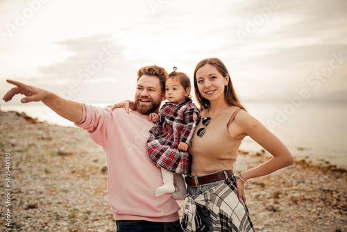 Young happy family spending their vacation on Italian Garda lake coast, bearded daddy pointing with finger, smiling mom, their little toddler girl in red checkered dress 