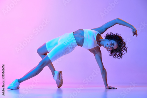 Flexibility of movements. Young woman with perfect fit body in white sportswear training against gradient pink blue background in neon. Sportive lifestyle, beauty, body care, fitness, health concept