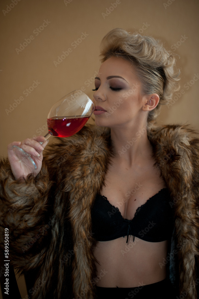 blonde model with pantyhose, glass of red wine ,black bra and fur coat posing provocatively on chair. portrait of sensual blonde, studio shot. Sensual female in black lingerie posing in vintage scene