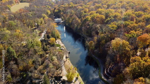 Beautiful panoramic autumn landscape park, many trees with yellow leaves, lake with fountain in center, architecture, big stones, paths walkways, people walking on sunny autumn day. aerial drone view