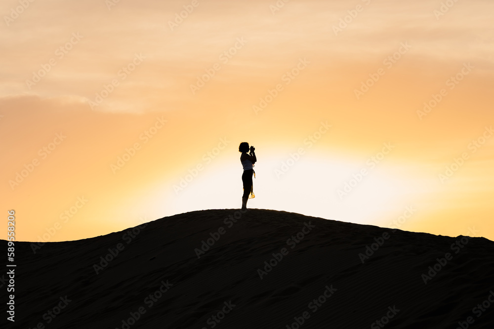 Silhouette of a beautiful girl that takes pictures from the top of a sand dune with a beautiful sunset in the background. Maspalomas, Grand Canaria, Canary Islands, Spain.