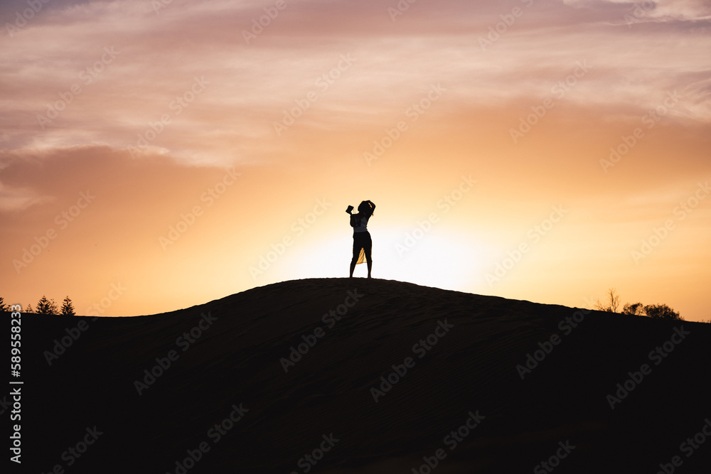 Silhouette of a beautiful girl that takes pictures from the top of a sand dune with a beautiful sunset in the background. Maspalomas, Grand Canaria, Canary Islands, Spain.