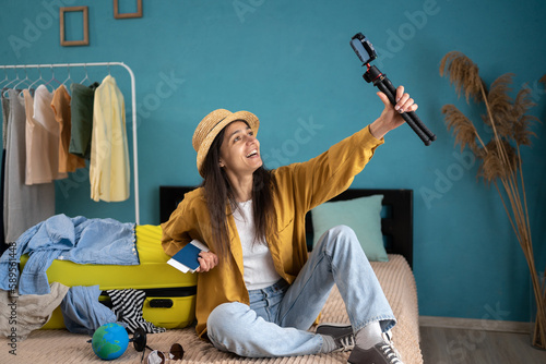 Social media vlogging concept. Young travel vlogger recording video content on smartphone, sitting on bed near the suitcase, speaking to her blog about journey. photo
