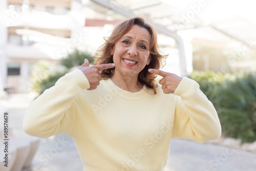 middle age woman smiling confidently pointing to own broad smile, positive, relaxed, satisfied attitude