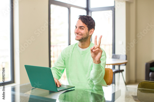 young hispanic man smiling and looking happy, carefree and positive, gesturing victory or peace with one hand. working at home