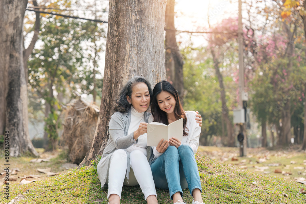 Lovely granddaughter and grandmother sitting under the tree in the green park, read a book together Lovely granddaughter and grandmother sitting under the tree in the green park, read a book together