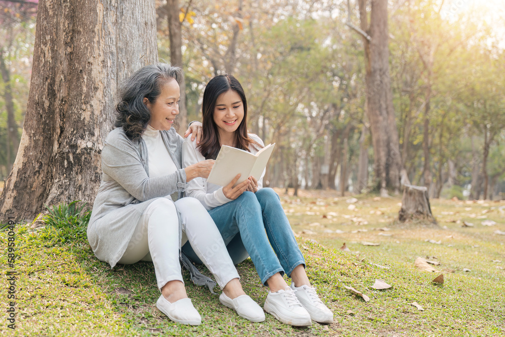 Lovely granddaughter and grandmother sitting under the tree in the green park, read a book together Lovely granddaughter and grandmother sitting under the tree in the green park, read a book together