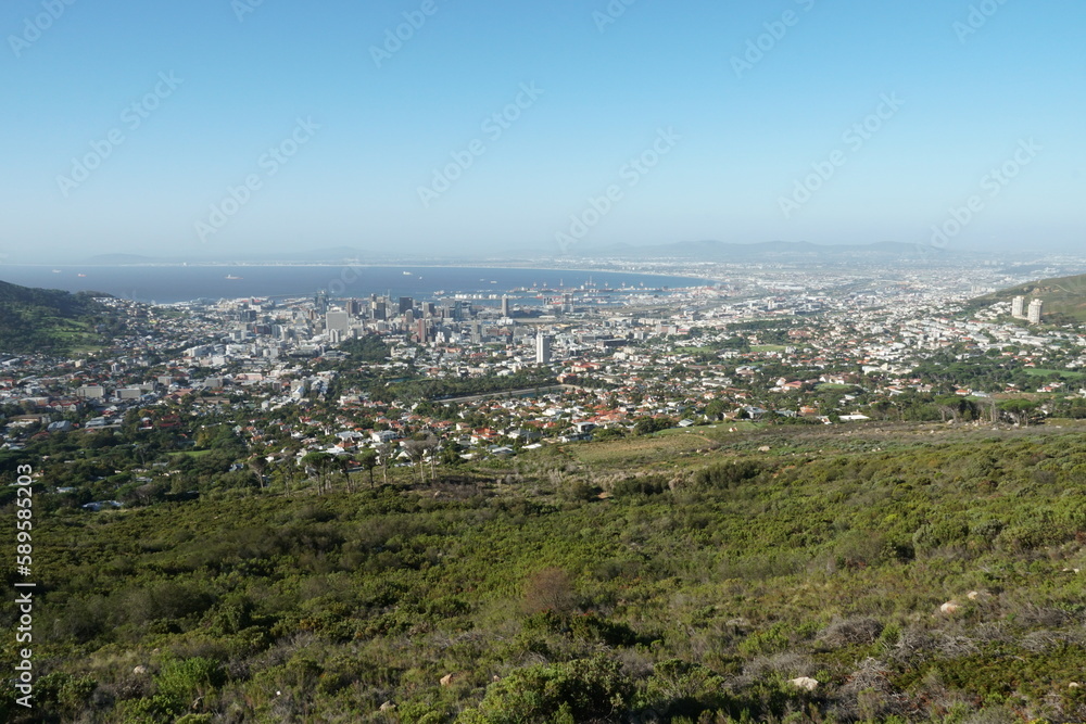 Panoramic view on Cape Town and Atlantic ocean from the foothill of Table Mountain overgrown by green grass and bushes. Townscape is from the base station of cable car which is running on the top.