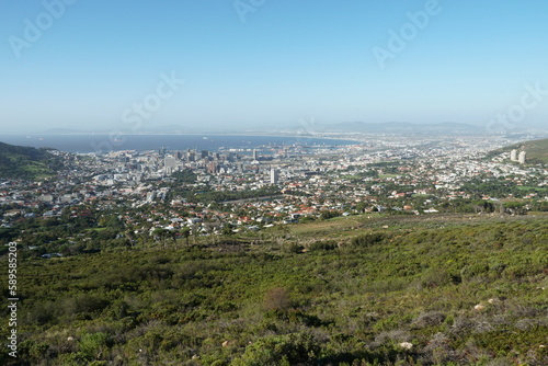 Panoramic view on Cape Town and Atlantic ocean from the foothill of Table Mountain overgrown by green grass and bushes. Townscape is from the base station of cable car which is running on the top.