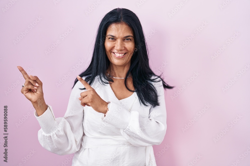 Mature hispanic woman standing over pink background smiling and looking at the camera pointing with two hands and fingers to the side. Mature hispanic woman standing over pink background smiling and looking at the camera pointing with two hands and fingers to the side.