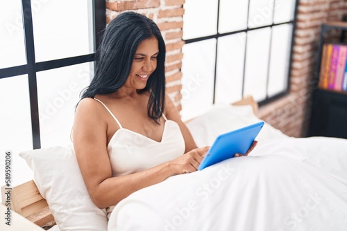 Middle age hispanic woman watching video on touchpad sitting on bed at bedroom