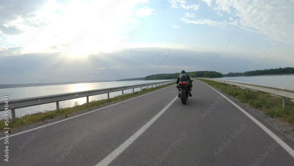 Biker riding on modern sport motorbike through road near lake. Motorcyclist racing motorcycle at dam route on summer day. Man driving bike through bridge of river. Concept of freedom and lifestyle Biker riding on modern sport motorbike through road near lake. Motorcyclist racing motorcycle at dam route on summer day. Man driving bike through bridge of river. Concept of freedom and lifestyle
