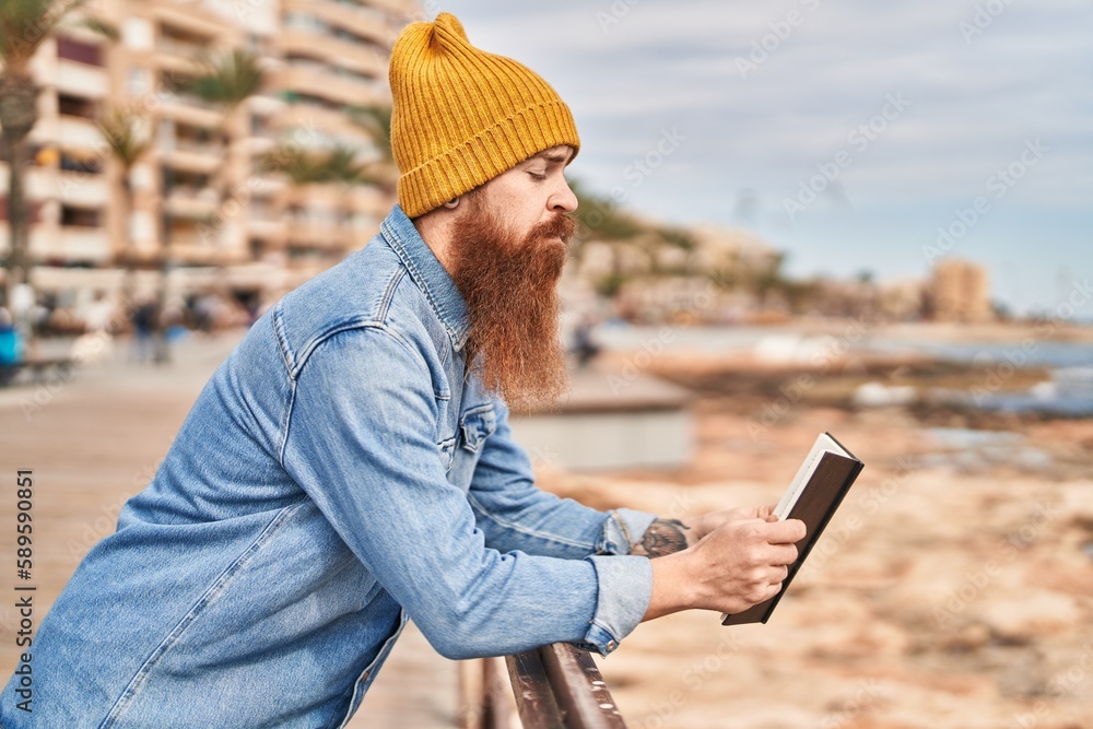 Young redhead man reading book with serious expression at seaside