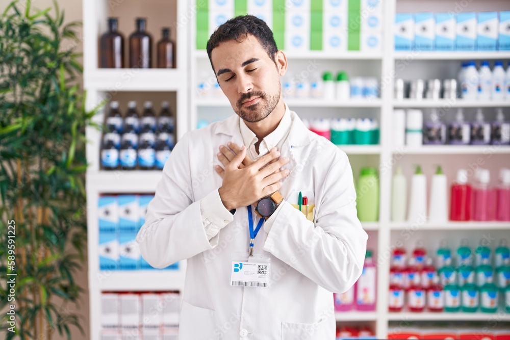 Handsome hispanic man working at pharmacy drugstore smiling with hands on chest with closed eyes and grateful gesture on face. health concept.