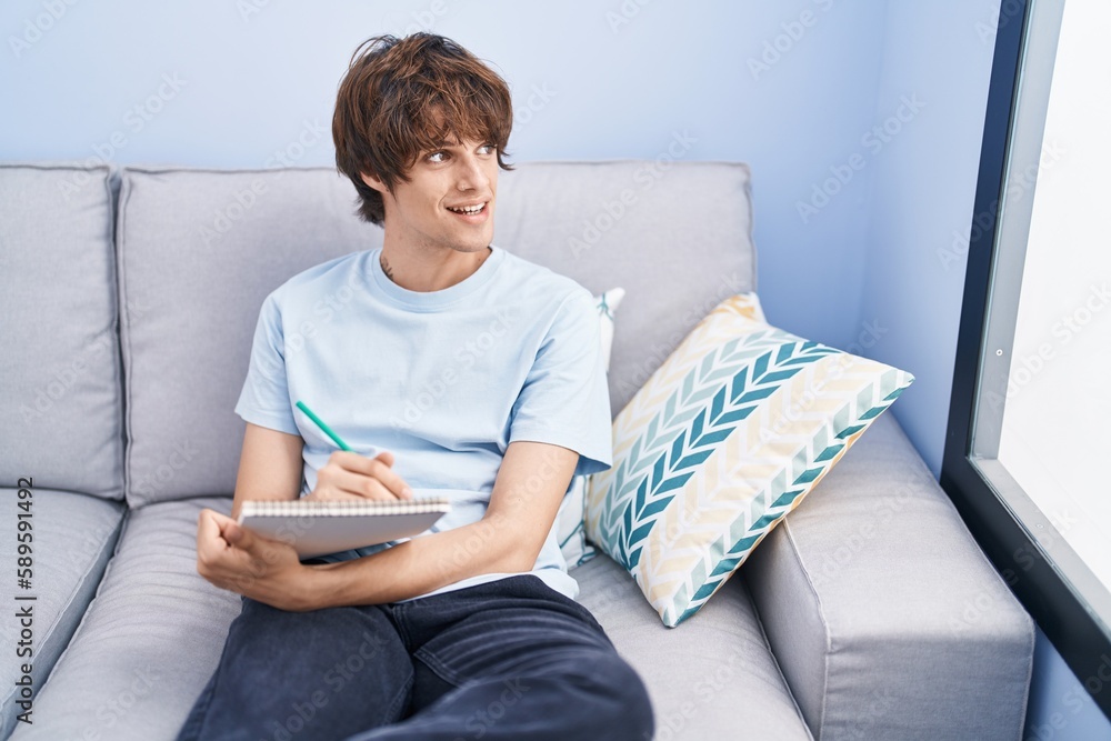 Young blond man writing on notebook sitting on sofa at home Young blond man writing on notebook sitting on sofa at home