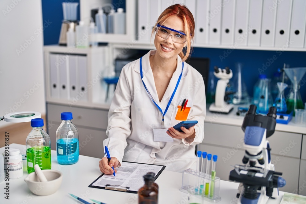 Young caucasian woman scientist using smartphone writing document at laboratory