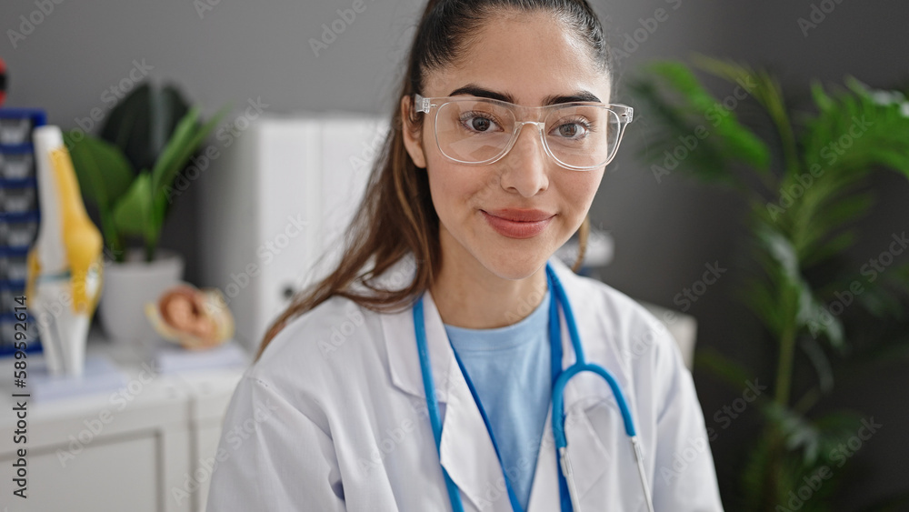 Young beautiful hispanic woman doctor smiling confident sitting on table at clinic