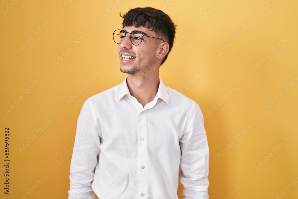 Young hispanic man standing over yellow background looking away to side with smile on face, natural expression. laughing confident. Young hispanic man standing over yellow background looking away to side with smile on face, natural expression. laughing confident.