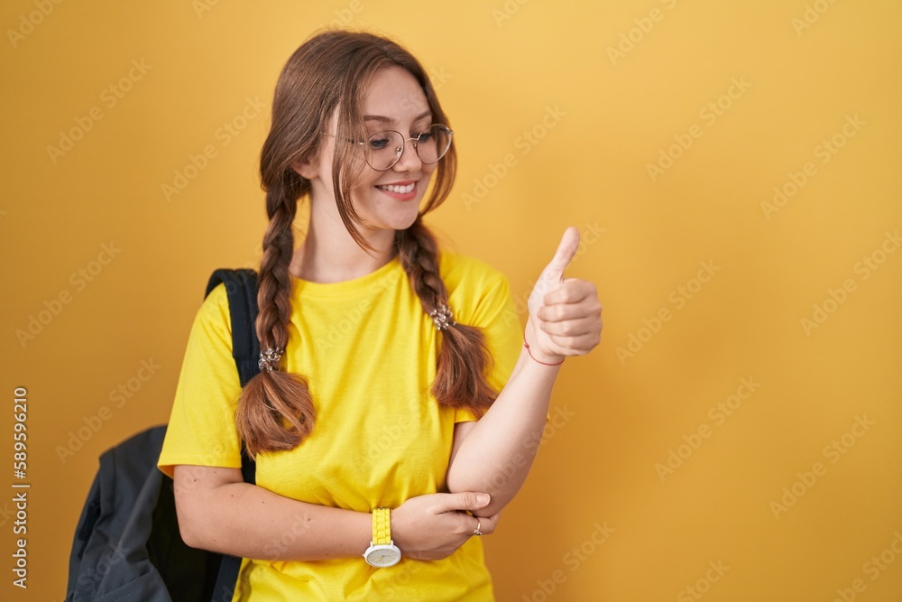 Young caucasian woman wearing student backpack over yellow background looking proud, smiling doing thumbs up gesture to the side Young caucasian woman wearing student backpack over yellow background looking proud, smiling doing thumbs up gesture to the side