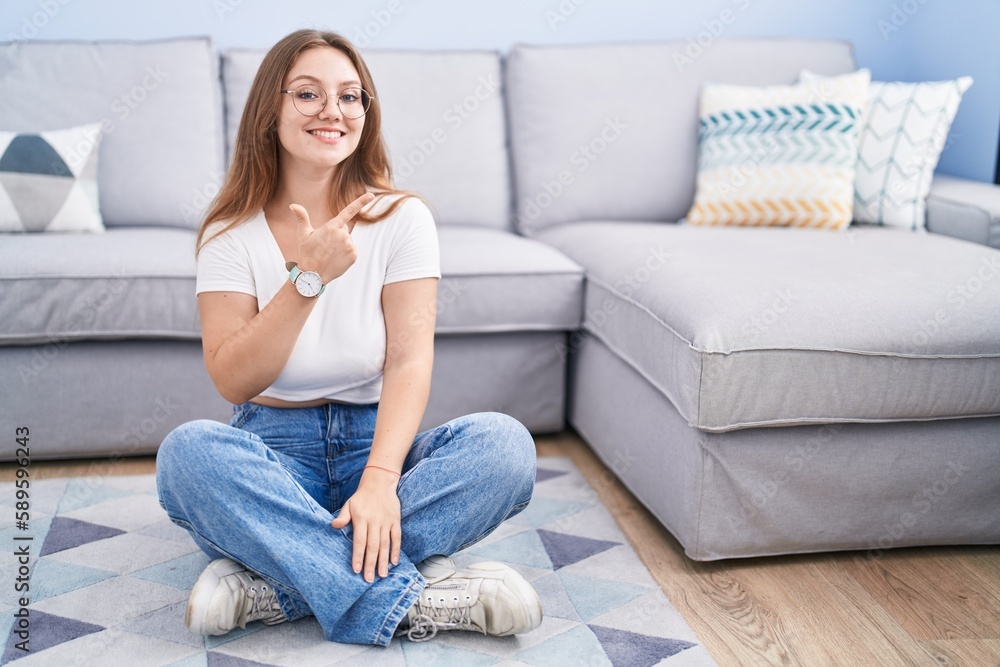Young caucasian woman sitting on the floor at the living room cheerful with a smile on face pointing with hand and finger up to the side with happy and natural expression Young caucasian woman sitting on the floor at the living room cheerful with a smile on face pointing with hand and finger up to the side with happy and natural expression