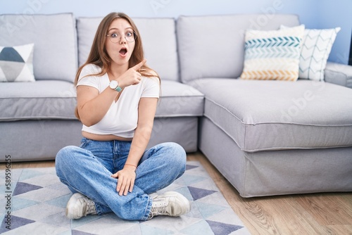 Young caucasian woman sitting on the floor at the living room surprised pointing with finger to the side, open mouth amazed expression.